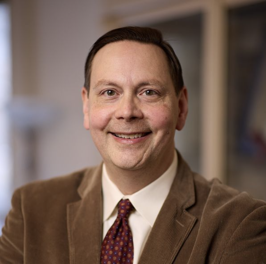 Photo of Steven Sandage, a Boston University School of Theology professor of psychology of religion and theology. He stands smiling in a white collared shirt, burgundy tie, and brown blazer. Blurred background shows a spacious office on a sunny day.